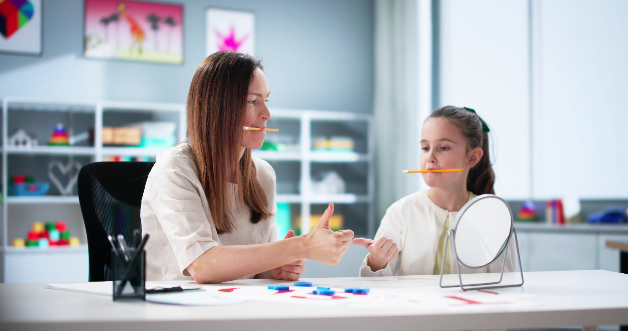Une femme et une jeune fille assises à une table, cheveux bruns, portent des crayons dans la bouche et se discutent autour d’activités pédagogiques, avec un miroir au burea et des éléments colorés sur la table.